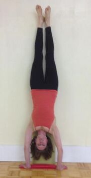 Picture of white woman doing a headstand. She has brown hair and is wearing a coral top and black tights.